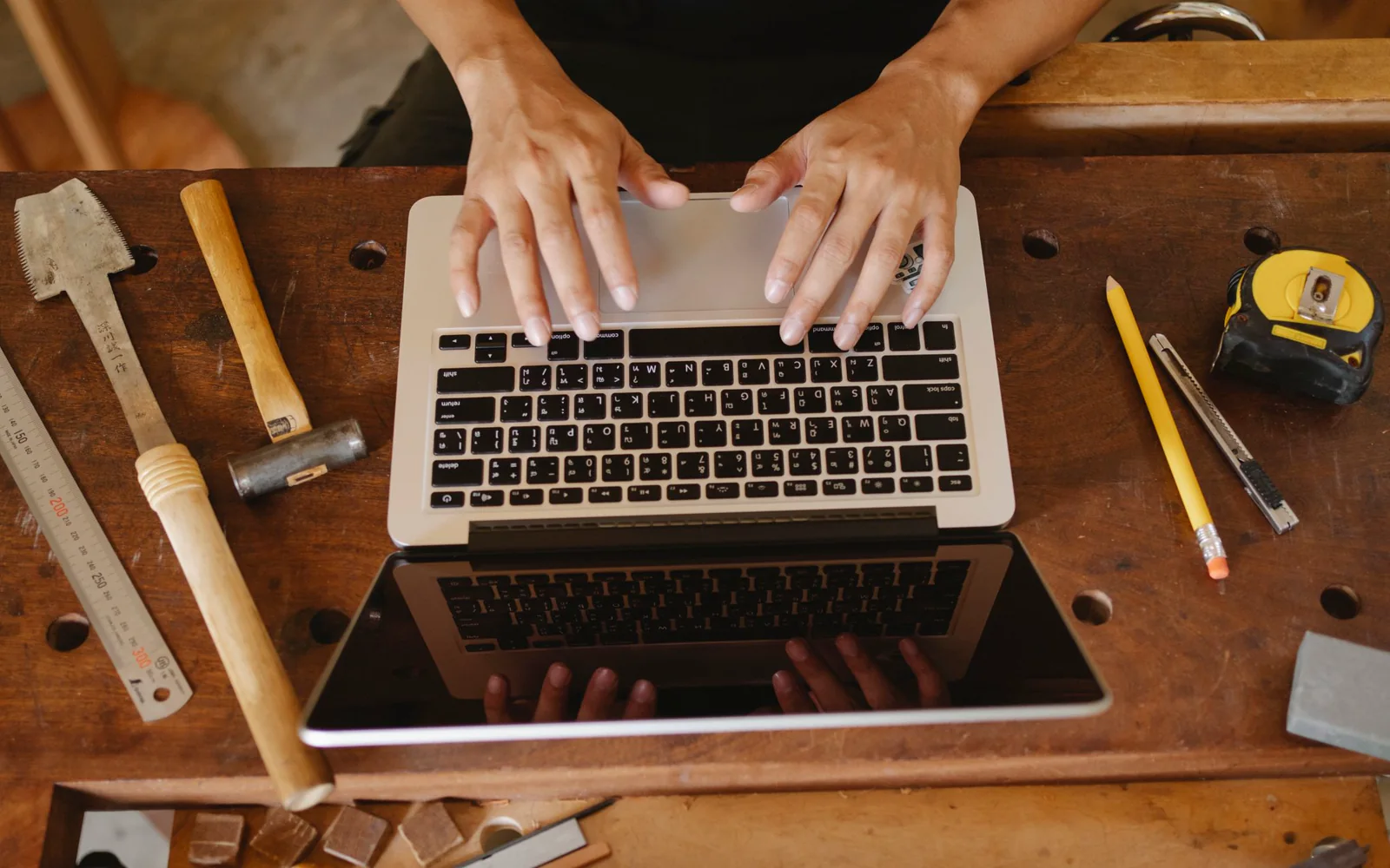 Hands using a laptop on a wooden workbench surrounded by measuring tools.